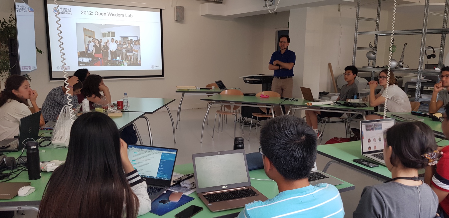 A professor teaching a master's class in the Solution Space, students following on laptops at the green tables
